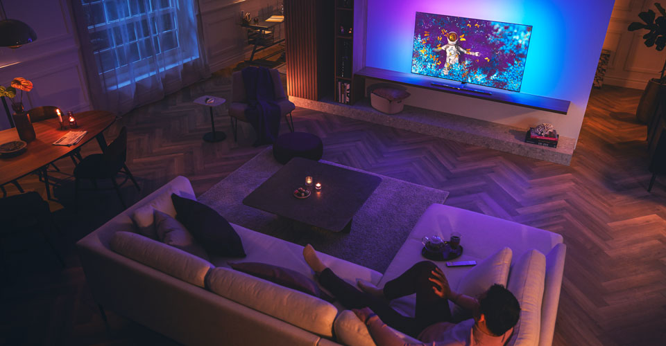 Mann slapper av p&aring; en sofa i en svakt opplyst stue og ser p&aring; en Philips Ambilight-TV